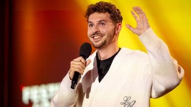 Fabian Lampert hält in seiner rechten Hand ein Mikrofon. Er trägt ein schwarzes T-Shirt und darüber eine weiße Jacke. Der Hintergrund ist rot und gelb beleuchtet. Er hebt seine linke Hand auf Kopfhöhe und zeigt seine Handfläche dabei nach vorne.