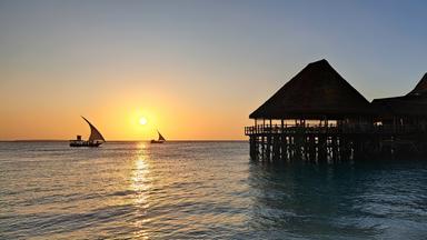 Abendstimmung am Meer; ein Boot segelt übers Meer, am Rand ein Steg mit einem hölzernen Haus. 