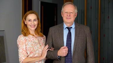 Eine Frau in gemusterter Bluse und ein Mann mit Krawatte und Jackett stehen eingehakt nebeneinander