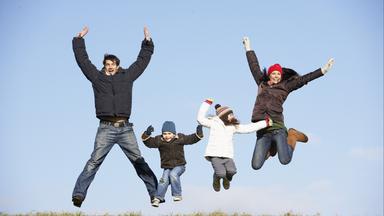 Ein Mann, eine Frau und zwei Kinder springen auf einer Wiese in die Luft, die Arme sind hochgereckt.