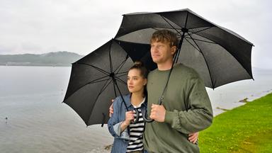 Ein Mann und eine Frau stehen mit Regenschirmen am Wasser.