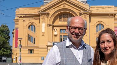 Zwei Personen stehen vor einem großen, gelben, historischen Theatergebäude im Freien. Der Mann trägt eine Brille, hat einen kahlen Kopf und grauen Bart. Er trägt ein helles Hemd und eine graue Weste und lächelt in die Kamera. Rechts steht eine Frau mit langen braunen Haaren, die eine helle Jacke und ein beigefarbenes Oberteil trägt. Auch sie lächelt freundlich. Im Hintergrund sind Bäume und Straßenschilder zu sehen, der Himmel ist klar und blau, es ist ein sonniger Tag.
