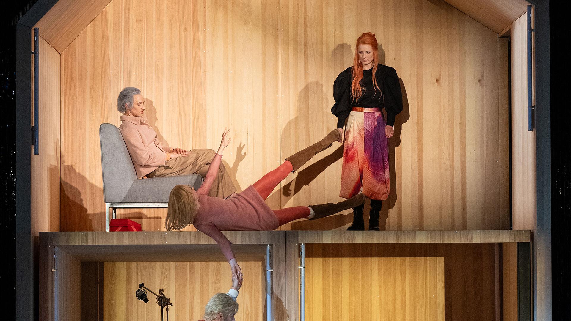 Bühnenszene: In einer Art offenem Puppenhaus mit zwei Stockwerken sitzen und stehen Menschen.