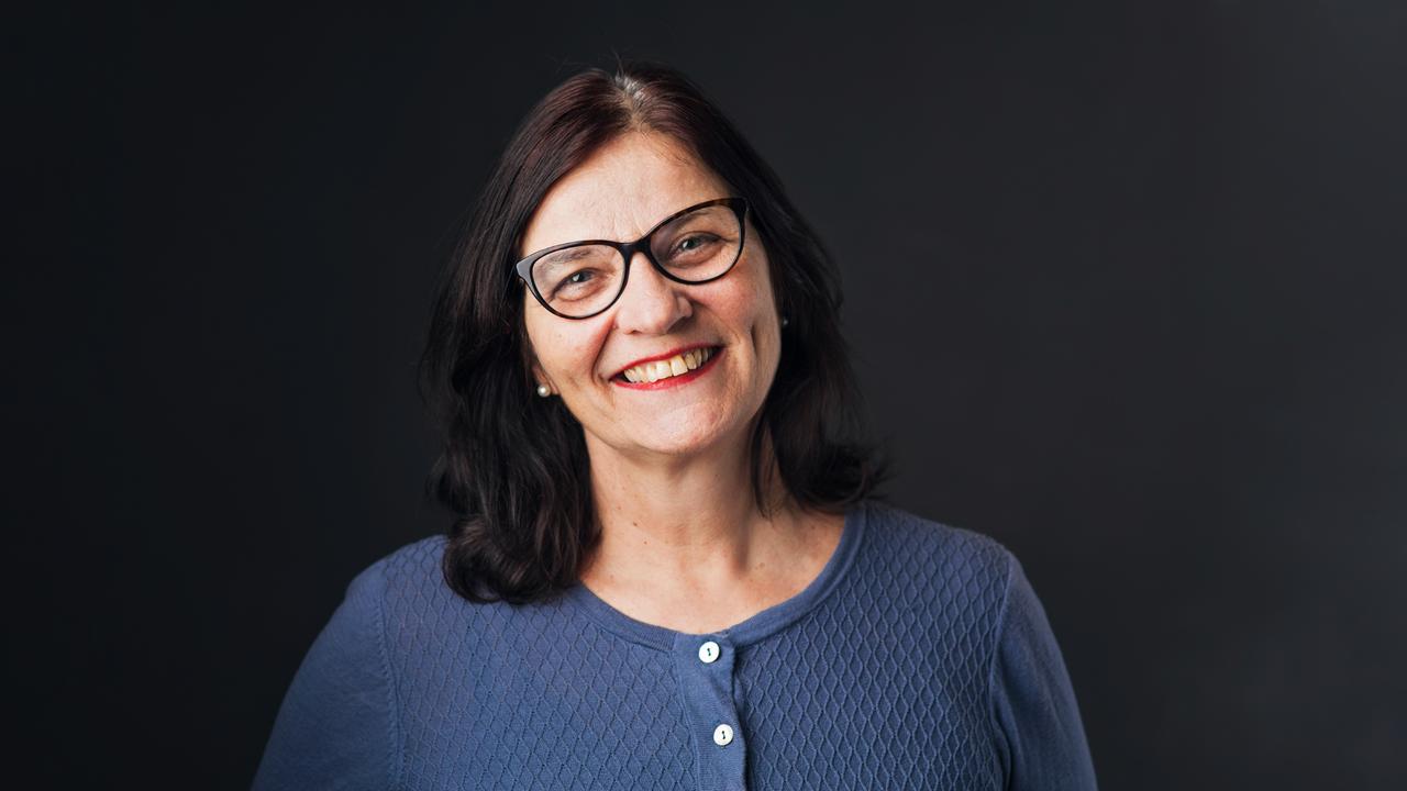 Porträt einer Frau vor dunklem Hintergrund. Sie trägt ein blaues Oberteil mit Knopfleiste und hat schulterlanges, dunkles Haar. 