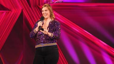 Fee Brembeck steht in der Mitte des Bildes. Sie streckt ihre linke Hand nach vorne. In der rechten Hand hält sie ein Mikrofon. Die Bühne ist rot beleuchtet. Fee Brembeck trägt eine glitzernde Jacke