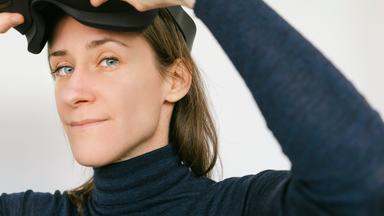 Das Bild zeigt eine Frau mit mittellangen, braunen Haaren. Sie trägt ein enges, dunkles Oberteil mit einem hohen Kragen. Ihr Blick ist konzentriert, während sie ein VR-Headset in beiden Händen hält und dabei ist, es auf ihren Kopf zu setzen. Der Hintergrund ist hell und neutral, wodurch die Aufmerksamkeit auf die Frau und das Headset gelenkt wird. Die Umgebung ist schlicht, ohne auffällige Elemente. Es handelt sich um eine Momentaufnahme, die den Einsatz von Virtual-Reality-Technologie suggeriert.