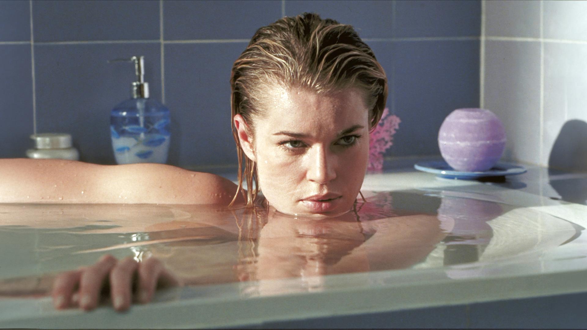 Femme Fatale: Laure (Rebecca Romijn) liegt in einer vollen Badewanne, ihr Gesicht knapp über der Wasseroberfläche, und blickt geheimnisvoll zur Seite.