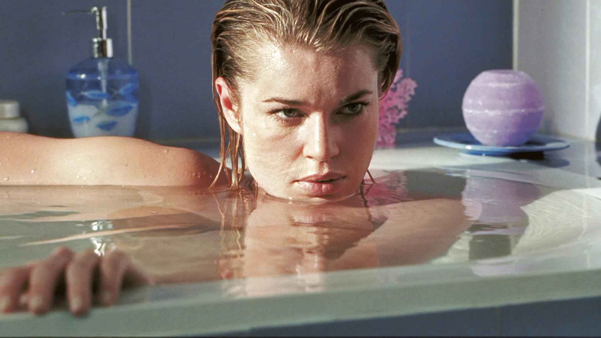 Femme Fatale: Laure (Rebecca Romijn) liegt in einer vollen Badewanne, ihr Gesicht knapp über der Wasseroberfläche, und blickt geheimnisvoll zur Seite.
