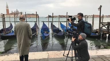 Ferdinand von Schirach steht mit dem Rücken zum Betrachter auf der linken Seite vor einem Kanal in Venedig. In der rechten Bildhälfte sind zwei Kameramänner beim Drehen. Am Ufer des Kanals liegen Gondeln und im Hintergrund sind man die Kulisse von Venedig.