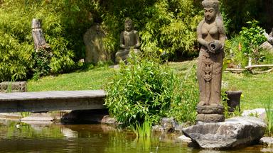 Das Bild zeigt einen Teil eines Gartens, der in der oststeirischen Gemeinde Floing angelegt wurde. Im Vordergrund ist eine Steinfigur einer weiblichen Gestalt zu sehen, die eine kleine Krug oder Vase hält. Diese Statue steht auf einem großen Stein, umgeben von dichtem Grün, das aus verschiedenen Pflanzen besteht.   Hinter der Statue erkennt man einen Teich, dessen Wasser ruhig ist und eine Reflexion der Umgebung zeigt. Auf der gegenüberliegenden Seite des Teiches ist eine Holzbrücke zu sehen, die über das Wasser führt und in den Garten hineinragt.   Im Hintergrund des Bildes sind weitere Pflanzen sowie eine weitere Statue zu erkennen, die eine meditative Pose einnimmt. Der Garten ist durch eine Vielfalt an Pflanzen geprägt, darunter auch Bambus. Die Atmosphäre wirkt friedlich und einladend, was den Garten zu einem Rückzugsort erscheinen lässt.