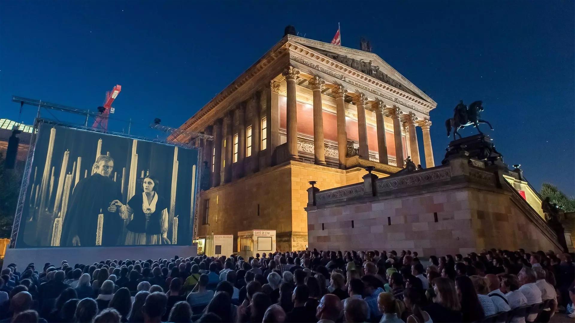 Man sieht ein historisches Gebäude, das aussieht wie ein griechischer Tempel, mit einer Kinoleinwand daneben, auf der ein Film vor Publikum gezeigt wird.