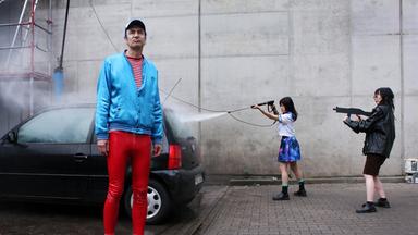 Ein Mann in roter Hose und mit einer blauen Jacke bekleidet, steht vor einem Auto. Rechts daneben steht eine Frau, die das Auto wäscht und eine Frau, die eine Maschinenpistole auf das Auto richtet.
