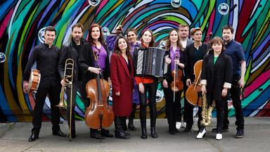 Ein Musikensemble mit Streich-, Blasinstrumenten und einem Akkordeon hat sich vor einer bunten Wand für ein Gruppenfoto versammelt.