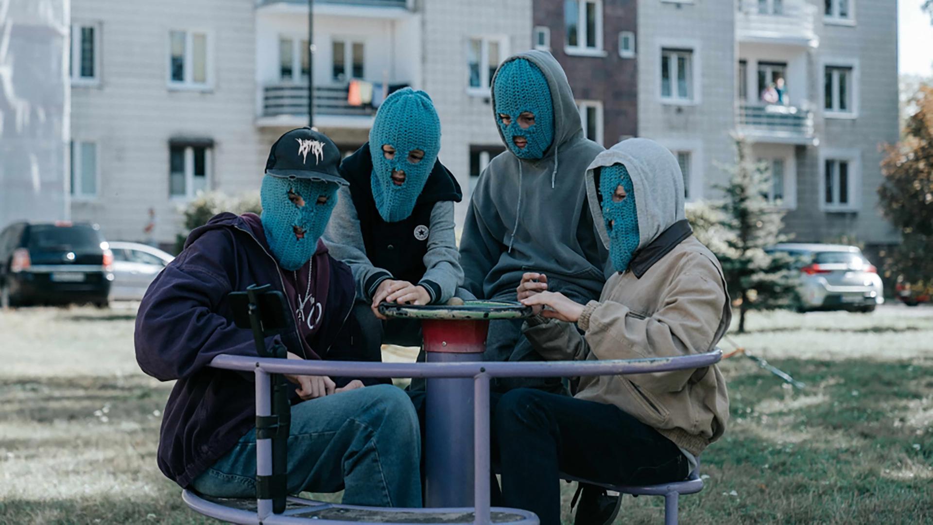 Vier junge Männer tragen blaue Strickmasken und sitzen auf einem Kinderkarussel.