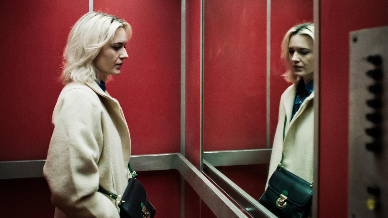 Man sieht eine Frau in einem Fahrstuhl, die sich in einem großen Spiegel betrachtet.