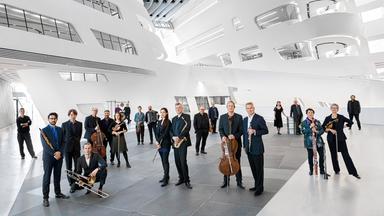 Menschen mit Musikinstrumenten stehen in einem futuristisch anmutenden Foyer.