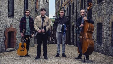 Vier Männer mit Musikinstrumenten stehen vor historischen Stein-Häusern.