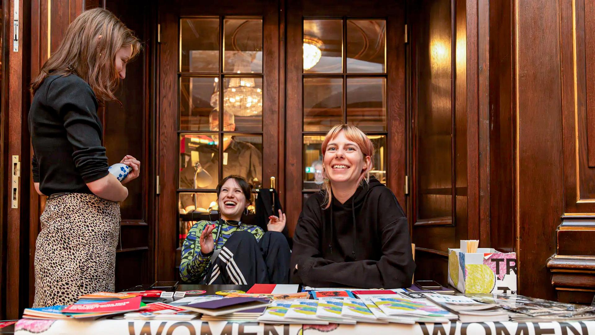 Drei Frauen stehen und sitzen an einem Informationsstand.
