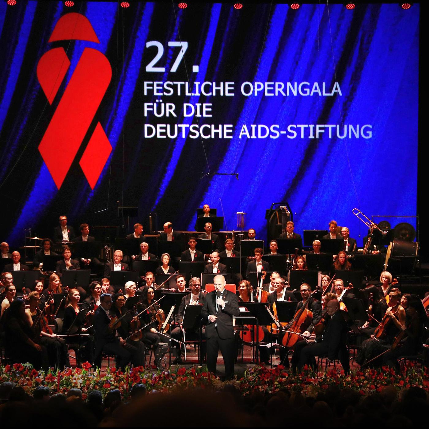 Blick auf ein Orchester, im Vordergrund spricht ein Mann in ein Mikrofon