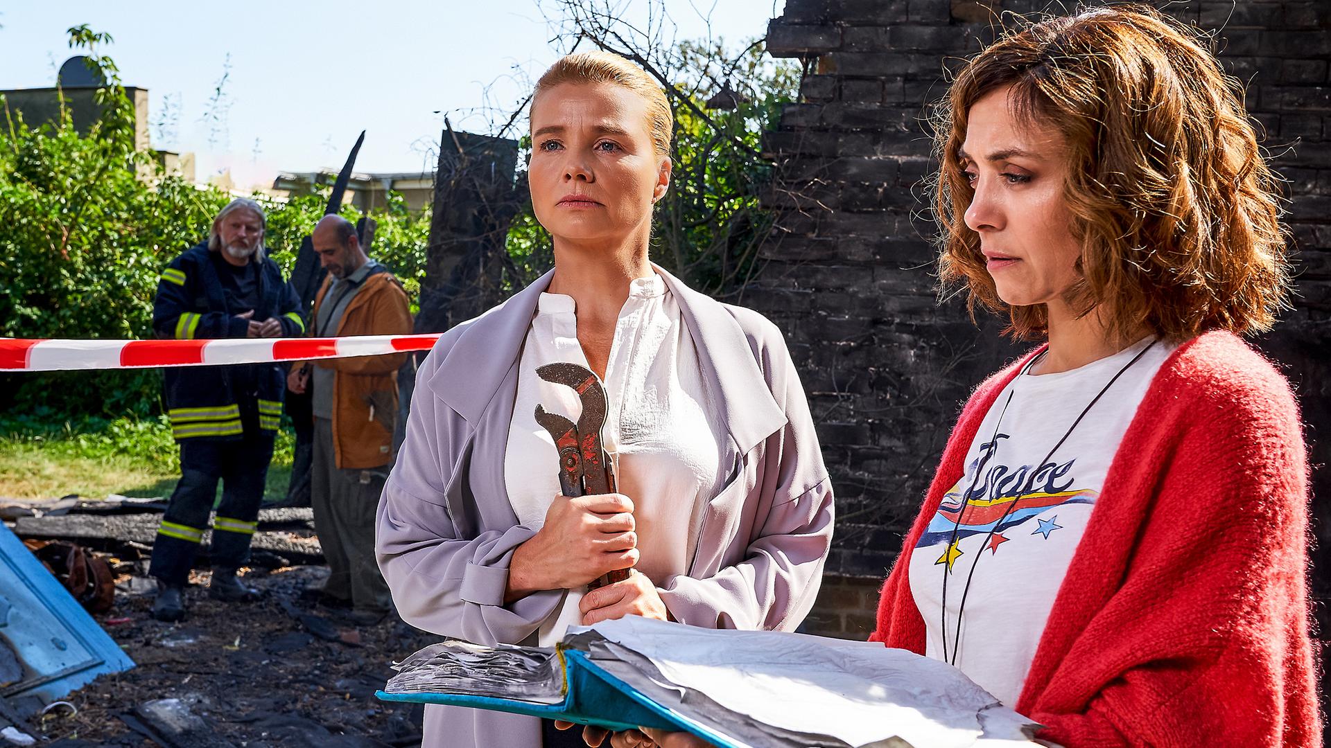 Im Hintergrund die Asche des zerstörten Hauses von Ella (Annette Frier) und Christina (Julia Richter), sowie ein Gutachter und ein Feuerwehrmann. Ella umklammert eine Zange, während Christina ein Fotoalbum in der Hand hält.