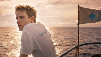 Ein junger Mann steht an der Reling eines Schiffes und schaut aufs offene Meer, das von einer tiefstehenden Sonne beleuchtet wird und schimmert.