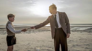 Filmszene: An einem Strand reicht ein Mann einem Jungen einen kleinen Gegenstand, der schlecht zu sehen ist. Beide tragen altmodische Kleidung aus der Nachkriegszeit. 