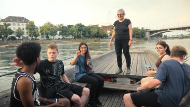 Eine Frau steht auf einem Boot, während  Kinder im Boot sitzen. Das Boot fährt auf einem Fluß.