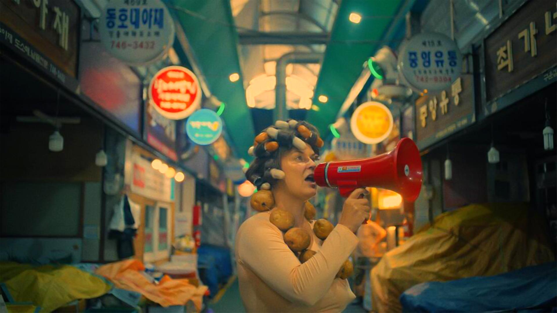 Eine Frau mit Lockenwicklern im Haar spricht durch ein Megafon, während sie durch einen koreanischen Markt geht.
