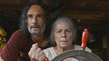 Ein Mann steht hinter einer älteren Frau die das Steuerrad eines Schiffes hält.