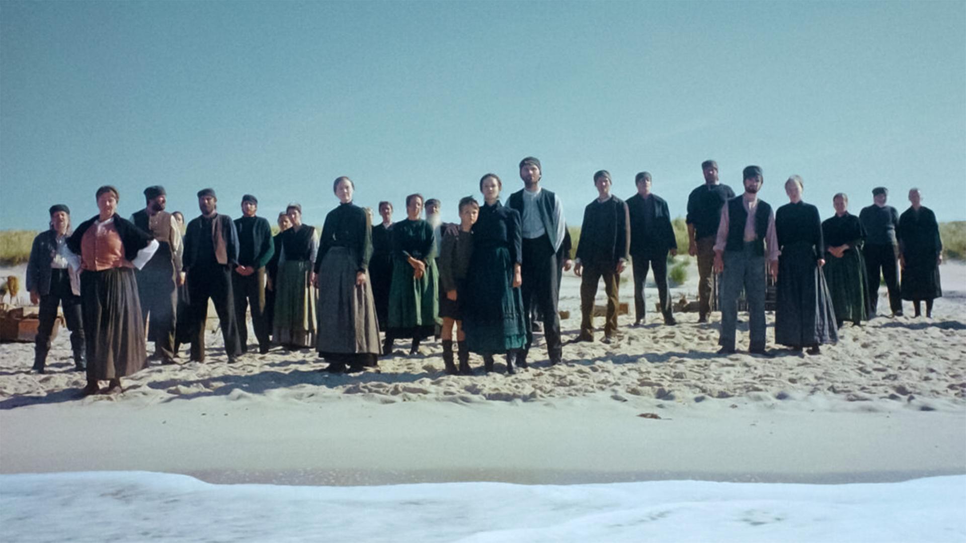 Eine Gruppe Menschen in historischer Bekleidung steht am Strand und schaut aufs Meer.