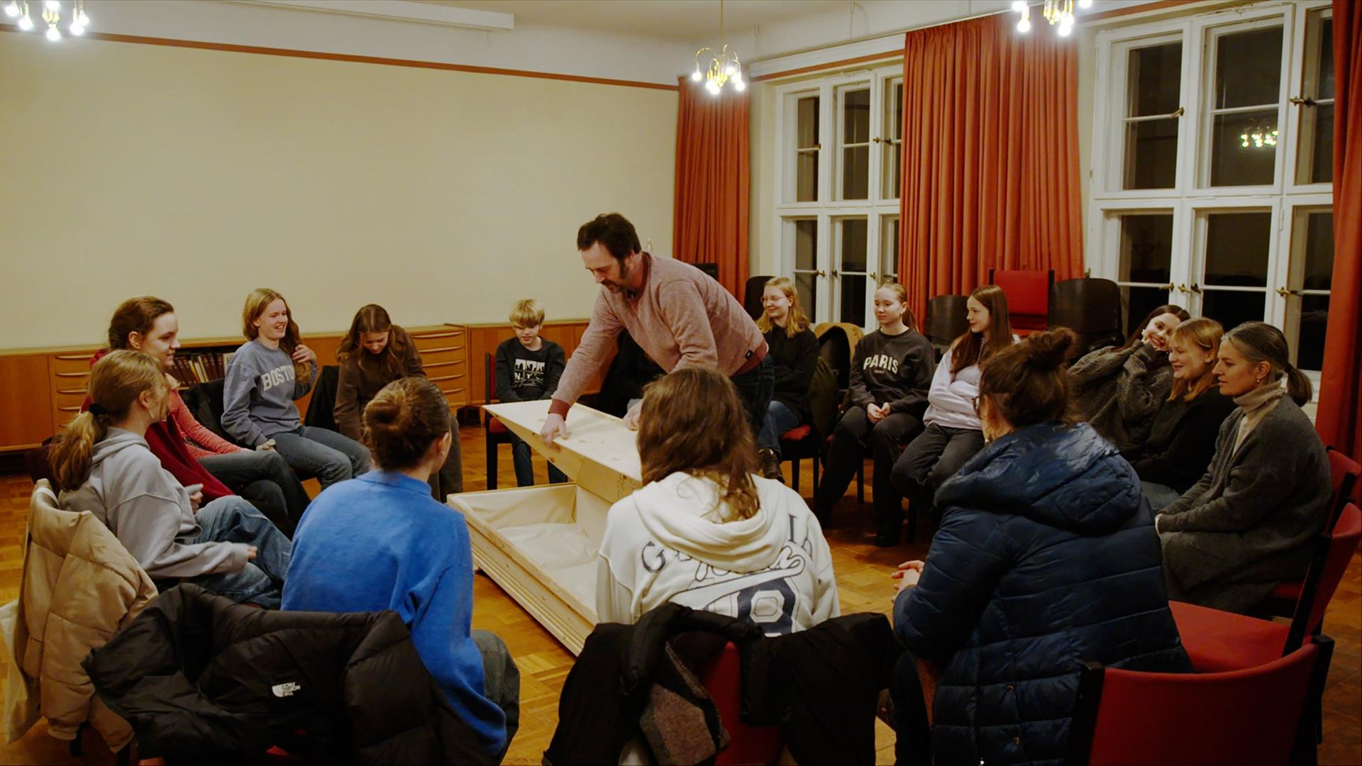Eine Schulergruppe sitzt in einem Stuhlkreis um einen Sarg, von dem ein Mann den Deckel abnimmt.