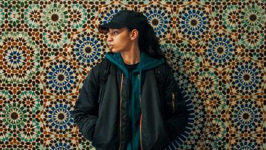 Eine junge Frau lehnt an einer bunten, Mosaik-Wand. Sie trägt eine schwarze Kappe, die dunklen Haare in einem Zopf und ein dunkles Outfit. Sie blickt zur Seite.