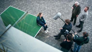 Ein junges Mädchen sitzt auf einer Tischtennisplatte. Vor ihr stehen fünf Männer mit Kamera und Mikrofonen, die sie zu interviewen scheinen.