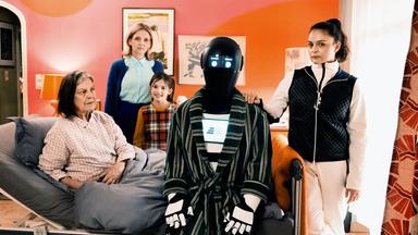 Eine ältere Frau sitzt in einem Bett, umgeben von zwei anderen Frauen, einem Mädchen und einem Roboter.