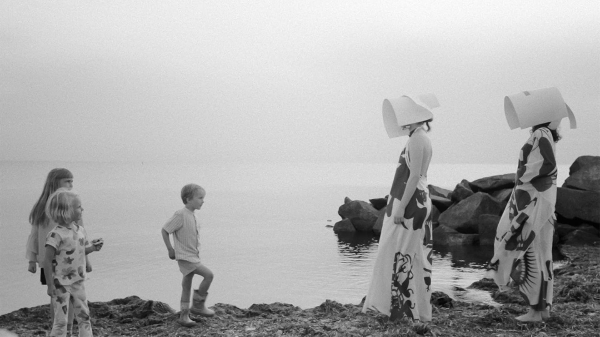 Auf einer Schwarz-Weiß-Fotografie sieht man drei Kinder und zwei Foto-Models in futuristischer Bekleidung am Strand.