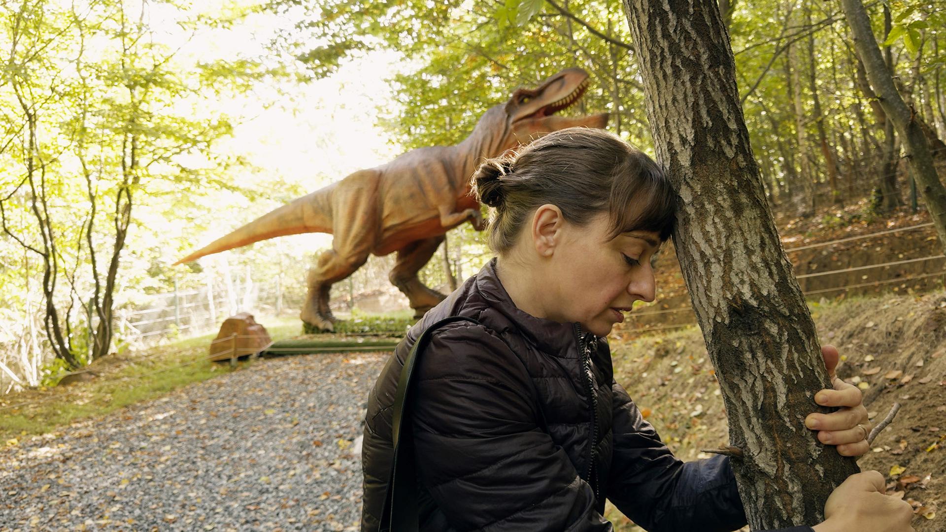 Eine Frau lehnt ihren Kopf an einen Baum und im Hintergrund steht ein Dinosaurier.