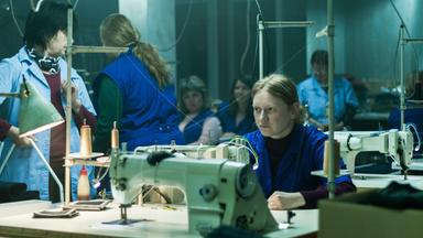 Eine blonde Frau in einem blauen Kittel sitzt in einer Werkshalle an einer Nähmaschine und schaut nachdenklich in die Ferne.