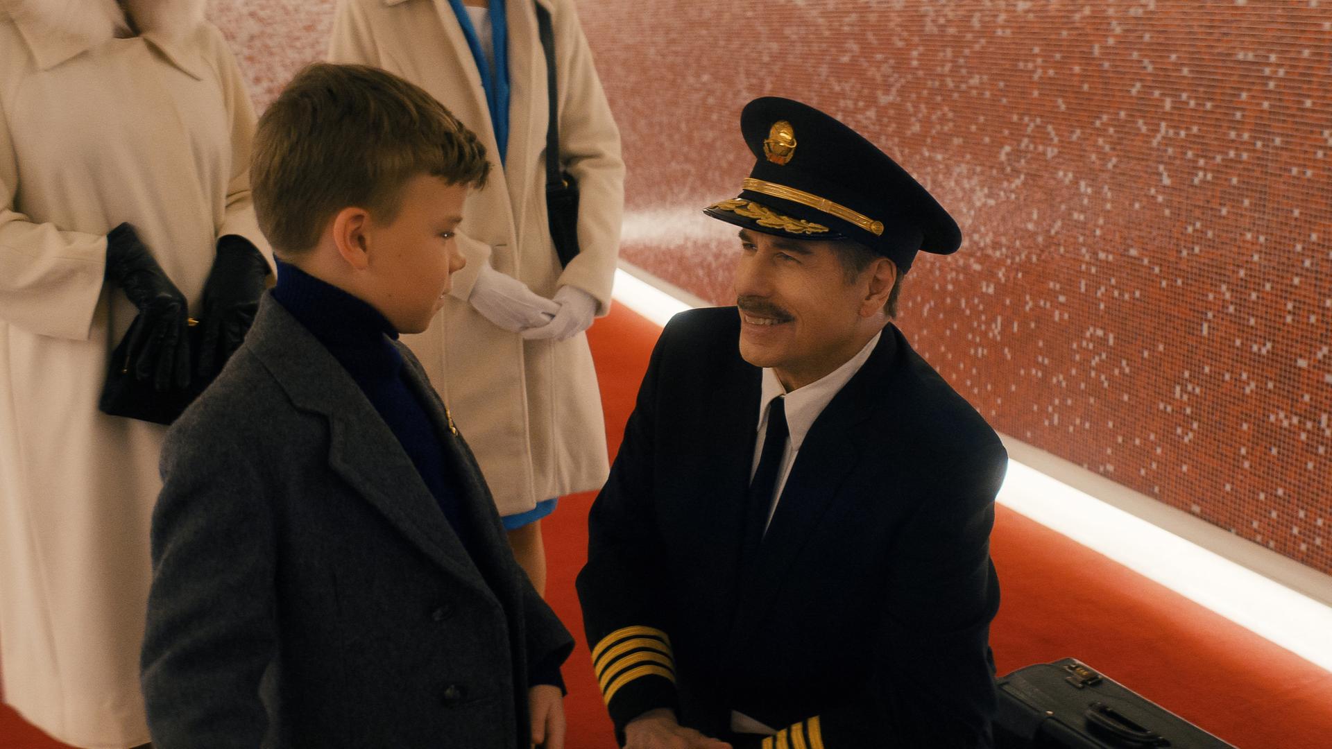 Ein Flugkapitän in Uniform kniet sich nieder, um mit einem kleinen Jungen in einer altmodischen Anzugjacke zu sprechen.
