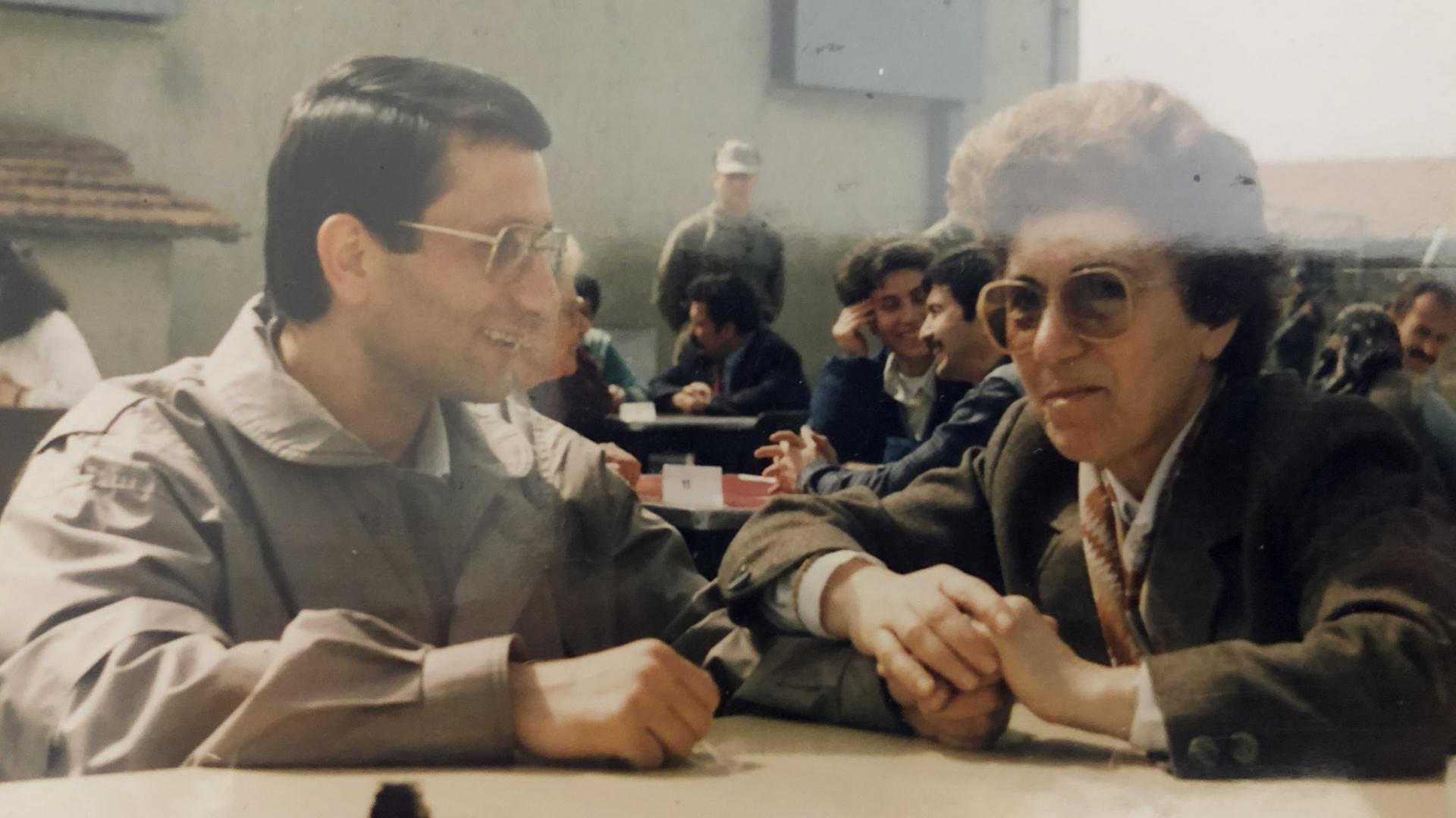 Zwei Personen, die Brille tragen, sitzen an einem Tisch und unterhalten sich. Sie tragen Kleidung im Stil der 1970er Jahre.