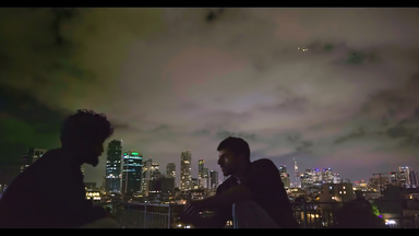 Zwei Männer sitzen abends vor der Skyline von Tel Aviv mit Hochhäusern. Sie sind nur als dunkle Silhouetten zu sehen.