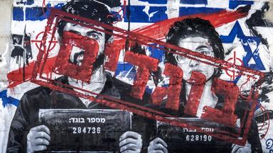 Auf einem Mural sind die Porträts eines Mannes und einer Frau zu sehen, die wie bei Verbrecherfotos Schilder mit Nummern tragen. Die Gesichter wurden mit hebräischer Schrift übermalt.