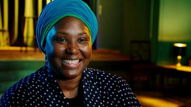 Man sieht eine schwarze Frau, die eine gepunktete Bluse trägt und einen blauen Turban auf dem Kopf.