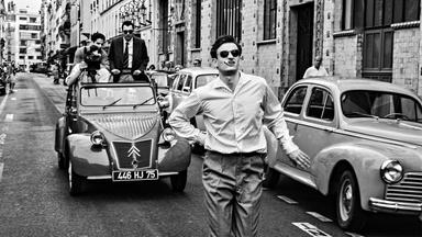 Das Schwarz-Weiß-Bild zeigt einen jungen Mann in einem Hemd, der mit Zigarette im Mund und Sonnenbrille zwischen Autos aus den 1950er Jahren auf einer Straße läuft. Hinter ihm auf der Straße fährt eine Cabrio-Ente mit offenem Dach. Drei Leute stehen in dem Auto, eine Person hat eine Kamera und filmt den Mann auf der Straße von hinten.