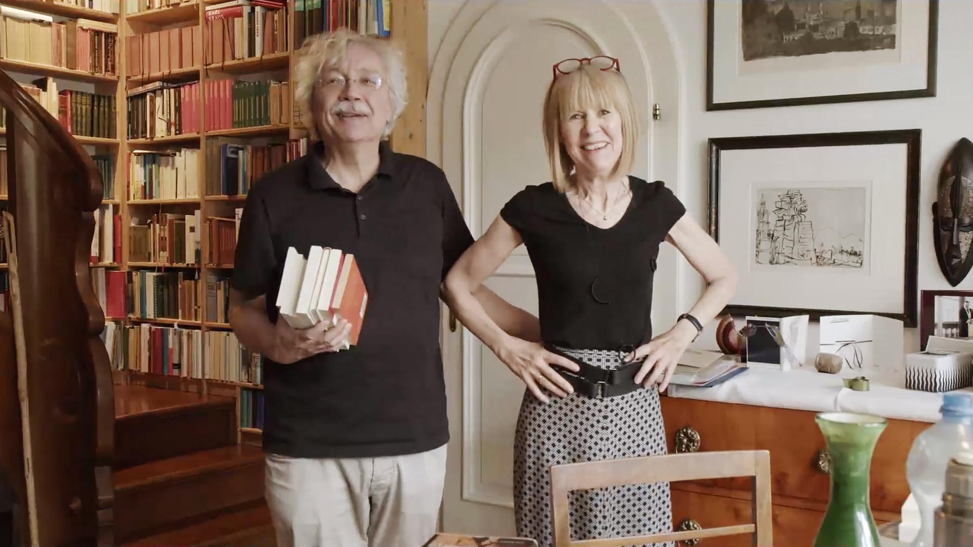 Ein Mann und eine Frau stehen in einem Raum mit Bücherregal im Hintergrund.