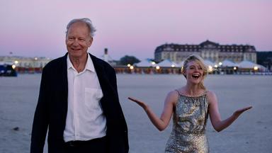 Stellan Skarsgård steht neben Elle Fanning, die ein Glitzerkleid trägt. Hinter ihnen sieht man das einen Fluss oder das Meer und in der Ferne Häuser.