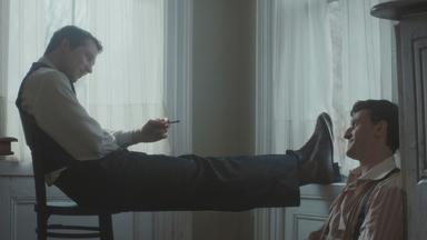 Ein junger Mann sitzt auf einem Stuhl, die Beine hochgelegt und eine Zigarette in der Hand, und schaut einen anderen jungen Mann an, der auf dem Boden an die Wand gelehnt sitzt. Die Männer lächeln einander an.