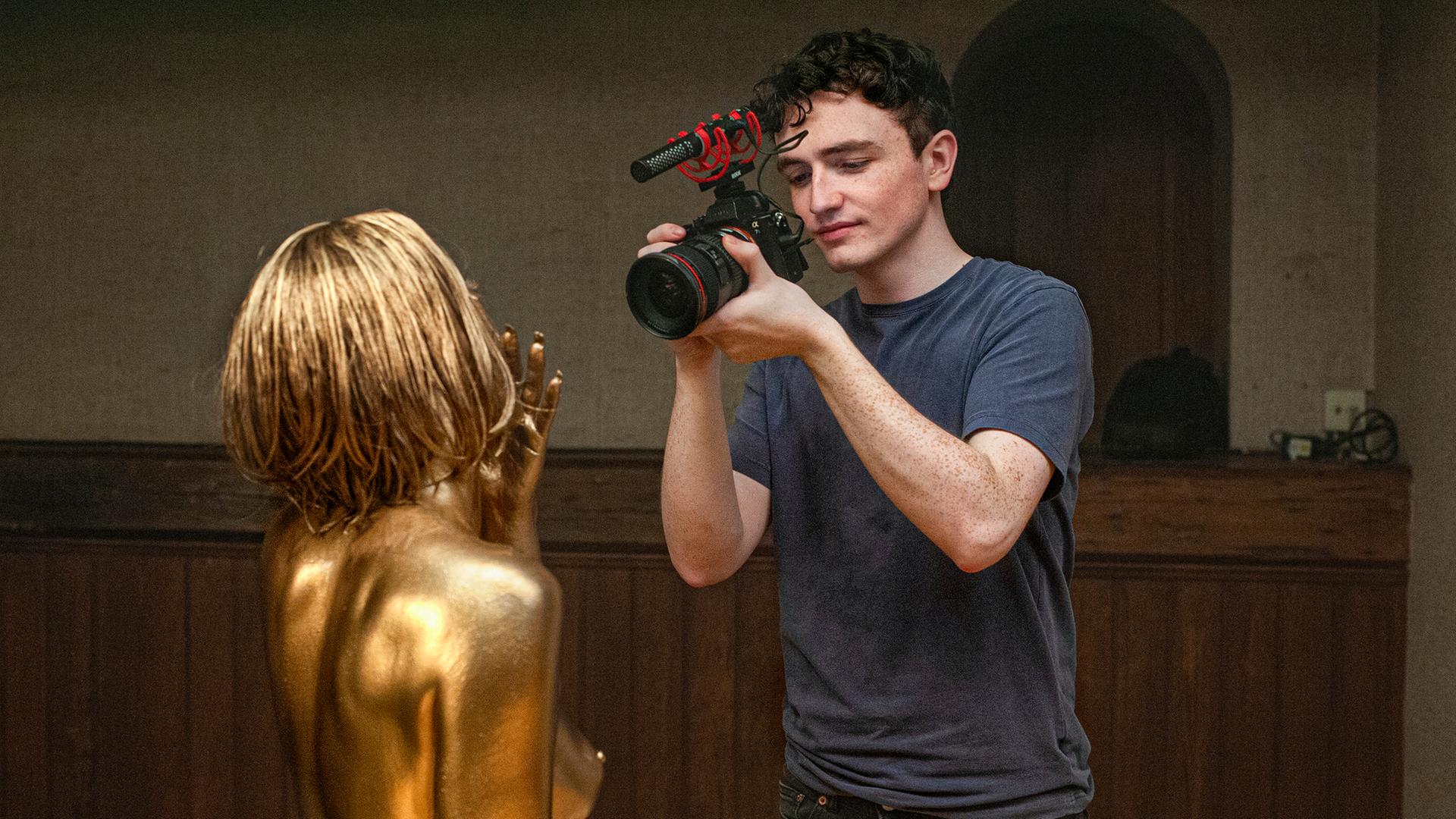 Rechts im Bild steht ein junger Mann, der eine Videokamera in der Hand hält und eine Frau filmt, deren Haare und Körper gold angemalt sind.