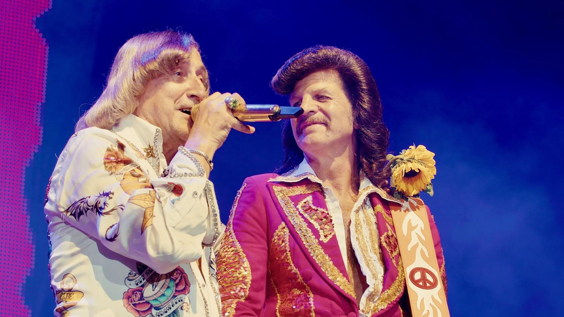 Zwei Männer in Kostümen der 1970er Jahre stehen auf der Bühne und singen.
