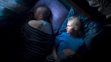 Eine junge Frau mit kurzen weißen Haaren liegt im Bett und betrachtet einen Mann mit kurzgeschorenen Haaren und Tattoos, der von ihr abgewendet neben ihr liegt und schläft.