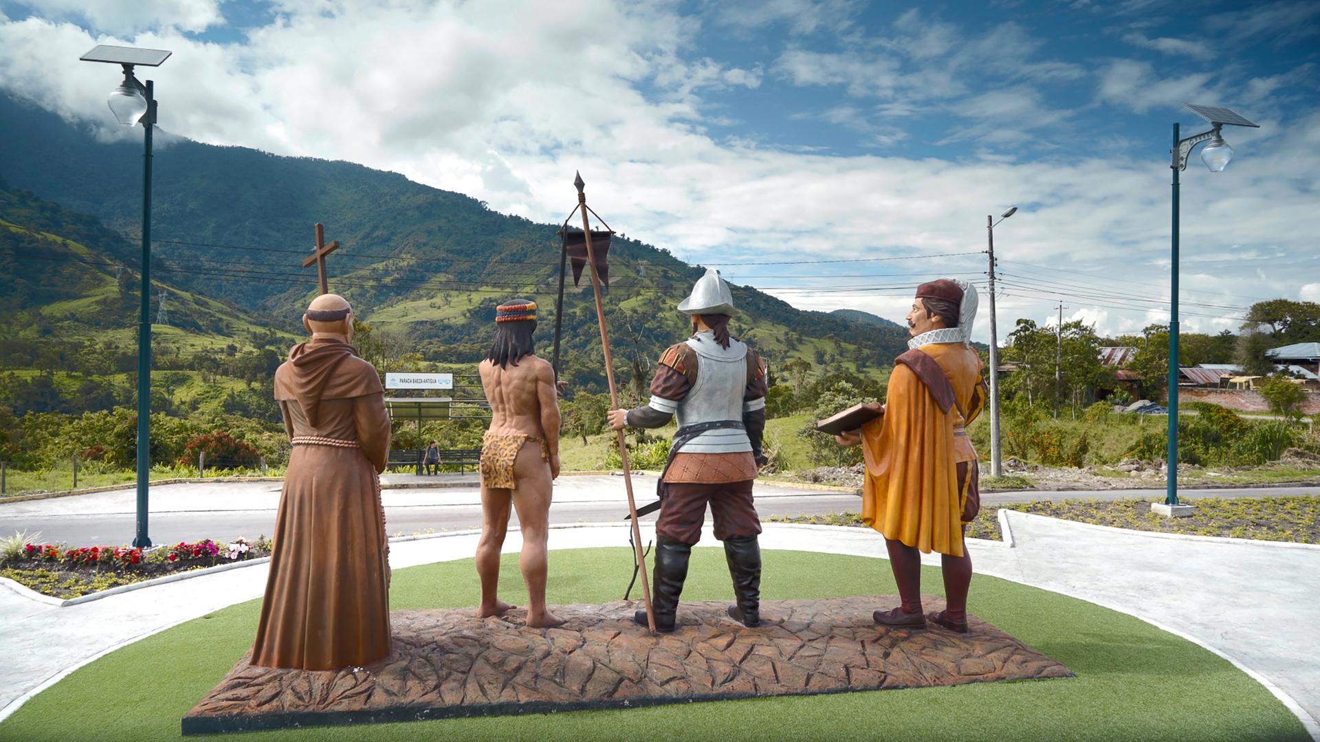 Vier Figuren stehen als Skulpturen in der Landschaft.